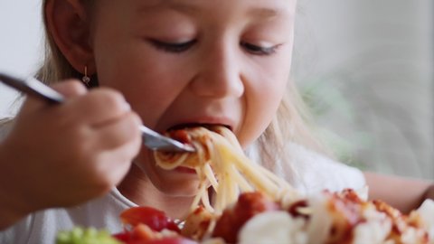 Close up portrait of a child girl eats spaghetti with salad, tomatoes, broccoli and red sauce in a light domestic room. A small child is twisting spaghetti on a fork and eating meal. 