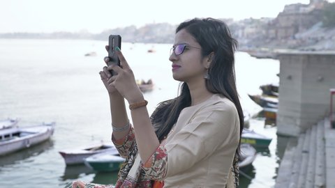 A young beautiful woman in traditional Salwar Kameez and wearing spectacles is taking images or photos with her cellphone camera near a riverbank.