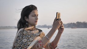 A beautiful smiling young woman wearing traditional Salwar Kameez making a video call on a cellphone with a river in the background during sunrise.