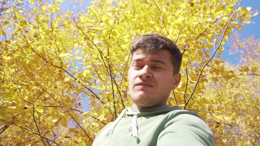 View of caucasian young nice-looking guy, wearing light-green hoody is standing under thin trees with yellow leaves somewhere in countryside on sunny Autumn day with blue sky.