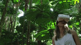 An attractive smiling Blonde Caucasian Girl wearing a white VR Headset explores a beautiful jungle scene with dappled light. Filmed on Red Camera, Slow Motion.