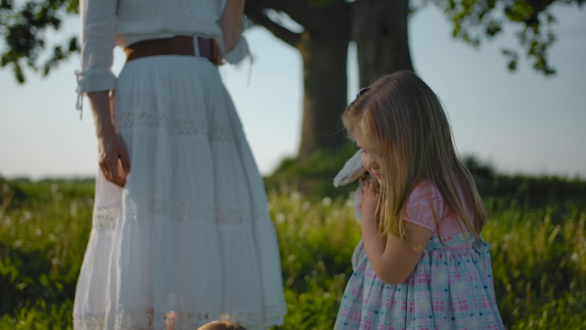 A happy daughter hugs a plush toy near her mom. She goes to her little sister smiling against the setting sun. Happy joyful carefree childhood next to my beloved mother and sister. Slow motion.