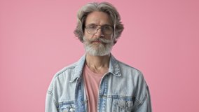 Cheerful elderly stylish bearded man with gray hair in denim jacket screaming and making winner gesture with hands while looking at the camera over pink background isolated