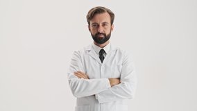 Pensive young bearded man doctor in white professional coat touching his beard and shaking his head negatively while looking at the camera over gray background isolated