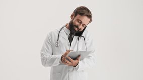 Happy young bearded man doctor in white professional coat with stethoscope working with medical history and talking on smartphone over gray background isolated