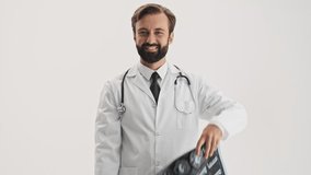 Happy young bearded man doctor in white professional coat with stethoscope smiling and looking attentively at two x-ray images over gray background isolated