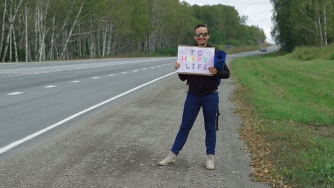 Cheerful Young Woman Hitchhiking and Holding a Cardboard Sign Inscription to Happy Life. Traveling, Lifestyle and People Concept