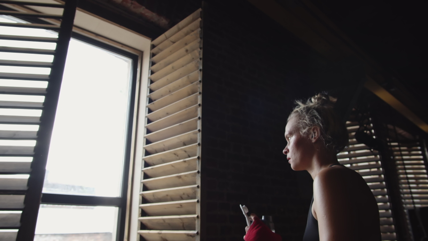 Medium shot of Caucasian young female MMA fighter with blonde hair standing inside boxing ring, resting and drinking water from sport bottle