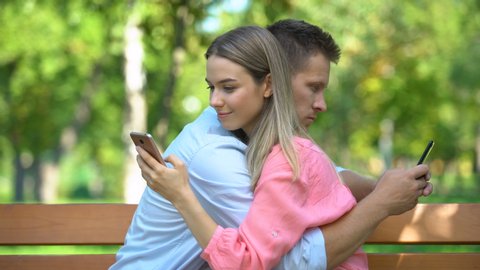 Young couple hugging at park, chatting on smartphones, lack of communication