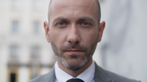Close up attractive middle aged bearded man in business suit looking into camera seriously in the city outdoors. Portrait of handsome serious male businessman standing on office porch