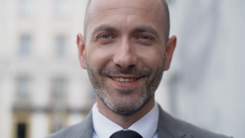 Close up attractive middle aged bearded man in business suit in a good mood looking into camera and smiling in the city outdoors. Portrait of handsome male businessman standing on office porch