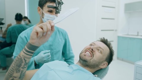 Young man patient and dentist with probe are checking teeth at mirror after dental treatment in dental office. Dentistry. Happy man patient and dentist