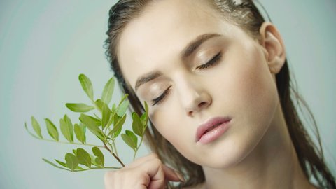 Face of young girl with clean smooth skin and small branch in hands. Natural and organic cosmetics, skin care, shampoo. Natural make-up. Girl holds leaves on her cheek. Opens eyes. Wet hair.