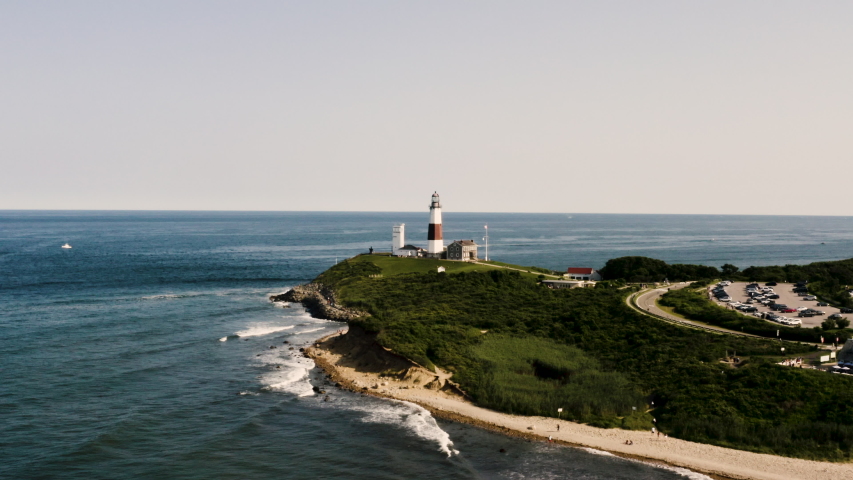 Stunning Aerial (Drone) Shot of Montauk Lighthouse in Long Island, New York surrounded by the blue sky, beach, seashore and the American flag Royalty-Free Stock Footage #1036613540