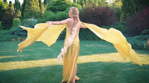 Joyful blonde woman with a long braid in a yellow dress. Cape train flies flutters in the wind. Rapunzel having fun is going round and spinning in the park. Background green trees landscaping lawn.
