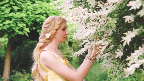 Attractive cute woman with braided hairstyle gently, she lovingly strokes a flowering tree. She sniffs the twigs and smiles softly. A fabulous image of a forest nymph in the Greek style. Royal garden
