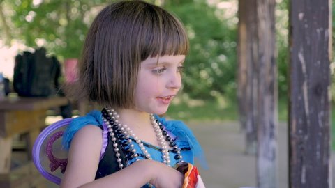 Young 4 - 5 year old girl with big blue eyes eating junk food cheese puffs wearing a fairy costume and Mardi Gras beaded necklaces