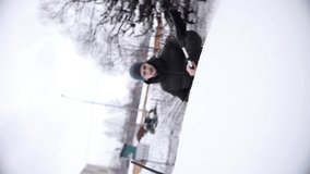 Mature man wearing blue ski cap and dark jacket, holding in hand black brush, removes snow from car top on cold winter day. Vertical video frame 9x16