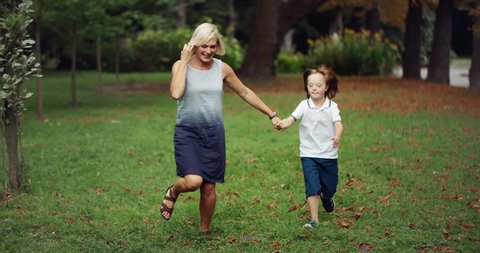 Smiling cute boy with Down syndrome holding mom's hand and running on green lawn in park, happy moments, childhood