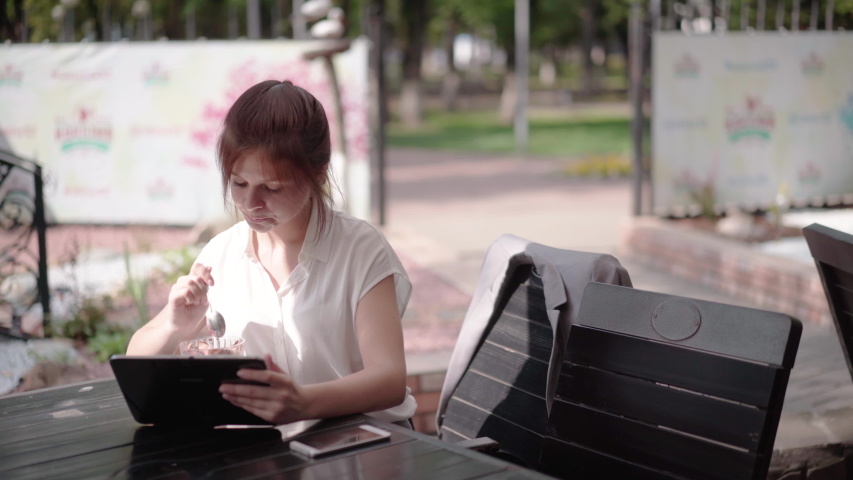 A beautiful girl sits in a cafe in the city center, uses the Internet by phone and at the same time eats ice cream
