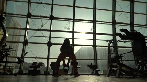 Bodybuilder girl on the background of large windows. She does a swing of a kettlebell down and up above her head.