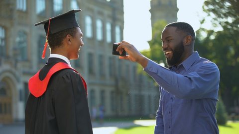 Black male taking photo of teenage son in graduation gown showing thumbs up