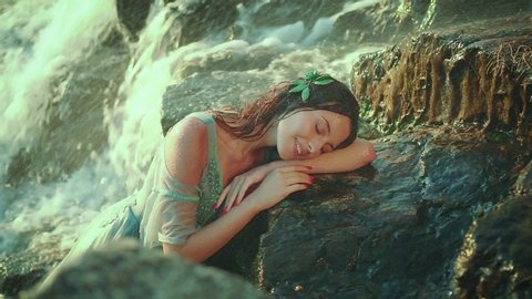 Young attractive woman in a wet water color dress lies on the stones. A tender, fragile, slender, river nymph sleeping beauty, resting near a tropical waterfall. Bright sunny day. Dreamy maiden.
