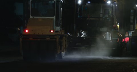 Road rollers silhouette working at night city on new road construction site. Truck laying fresh asphalt in town. Team of workers put hot asphalt on a street. Steam rising from hot asphalt surface.
