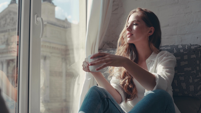 Gorgeous young woman in the house holding and drinking a cup of tea near the window smiling looking outside feel happy | Shutterstock HD Video #1036735622