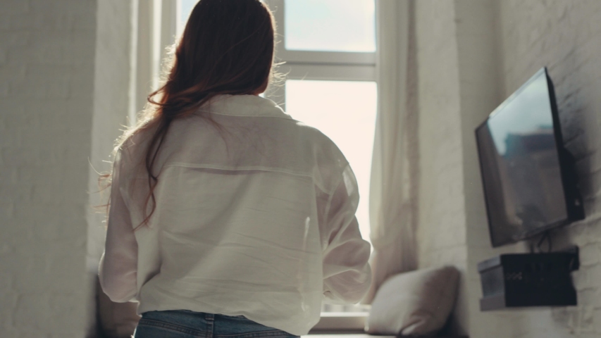 Slender young woman with curly red hair goes to the window in the apartments with a cup of tea and looking to the street