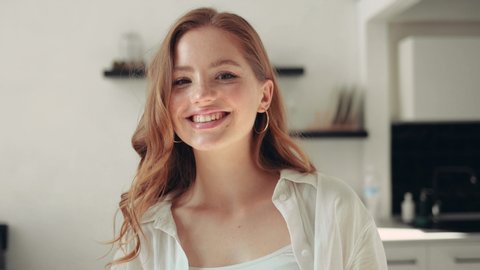 Close-up portrait of cute redhead girl with curly hair and freckles looking at camera smiling inside the room positive emotion
