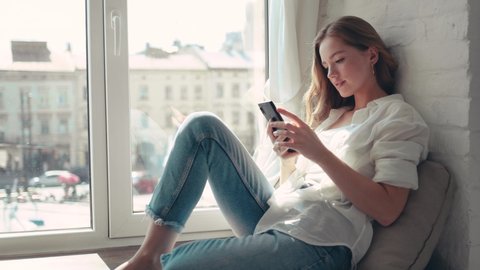 Charming young redhead woman with curly hair in the apartment looking through the window holding and checking a smartphone