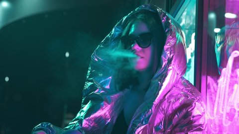 Millennial cool modern pretty girl smoking cigarette near glowing neon wall at night. Mysterious hipster. Beautiful stylish teen wearing shiny laser jacket and sunglasses. bad lifestyle