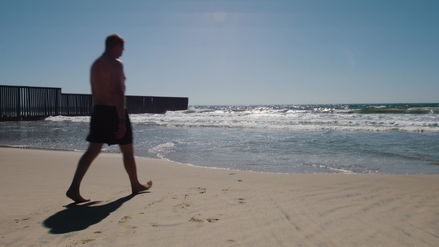 Adult Man walks towards Ocean along border wall between San Diego & Mexico