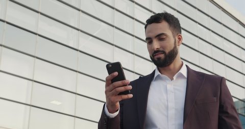 Well Dressed Young Businessman Holding Mobile Phone. Walking in the City. Checking his email. Reading new Messages. Looking Happy. Classical Outfit. Charming smile. Relax after Workday.