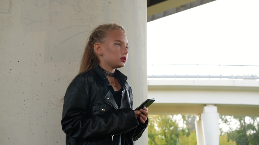 Frozen girl in black leather jacket trembling from cold under car bridge in city. Punk girl with heavy makeup face holding mobile phone on cold urban street