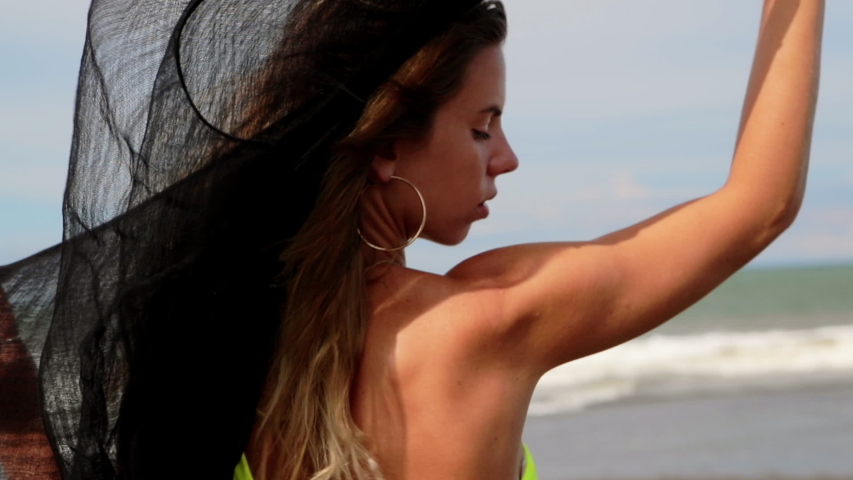 Beautiful young woman in neon yellow bikini posing to the camera with light transparent black fabric flying in the wind. Concept of fitness healthy active living. Slow motion. Sport and travel.