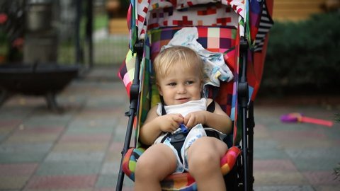 Portrait of adorable cute caucasian blond toddler boy sitting bright multicolored stroller and being rocked by mother during walk at backyard outdoors