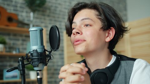 Close-up slow motion of happy teenager blogger speaking in microphone at home recording audio smiling enjoying activity. Youth culture and lifestyle concept.