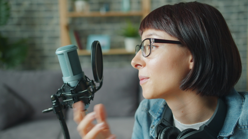 Close-up of pretty girl in glasses recording audio talking in microphone working in apartment alone. Modern technology, lifestyle and blogging concept. | Shutterstock HD Video #1036772099