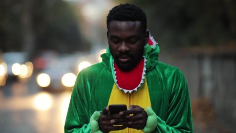 Portrait of thoughtful african guy sociable green funny dinosaur texting sms on smartphone standing on roadside in busy street.
