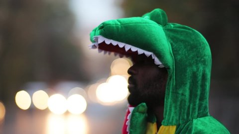 Handsome afro-american man in green fun dino costume staying outdoor looking serious. Busy traffic cityscape, blurred background.
