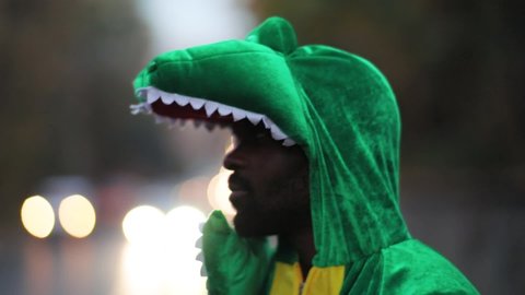 Funny afro-american actor playing lovely dinosaur wearing green costume standing smiling on busy street cityscape lights blurred background.