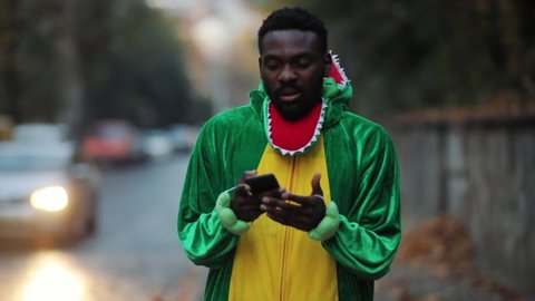 Funny color dinosaur young african man using smartphone thinking texting message in social network standing outdoor traffic street cityscape background.
