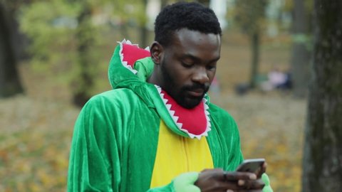 Portrait of attractive black african man in dinosaur costume using smartphone messaging thinking staying in the park on lovely autumn day.