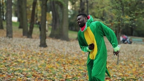 Young african man in dinosaur fun costume using modern smartphone excited with news cheering bending of stomach ache staying in natural city park.
