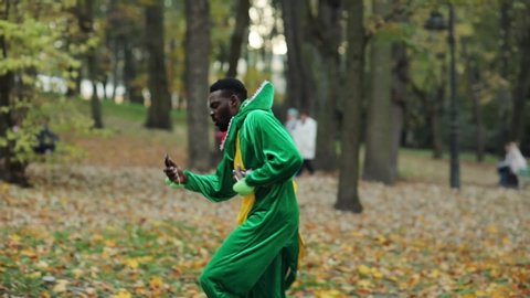 Young afro male dinosaur rejoicing good news using smartphone twisting of stomach pain need help staying in autumn city park.