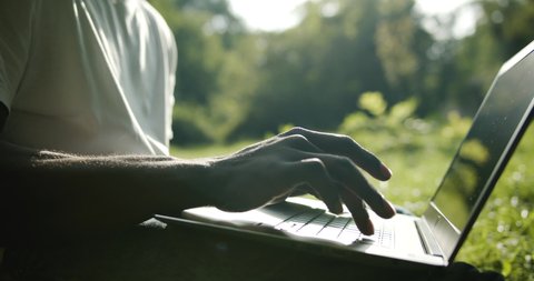 Young african-american male typing on laptop keyboard sitting on green grass in park, spending time outdoors