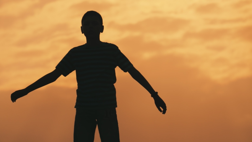 Impressive view of a sportive ten-years-old boy with a short haircut standing and waving his hands doing exercises in a relaxed way at orange sunset in summer.