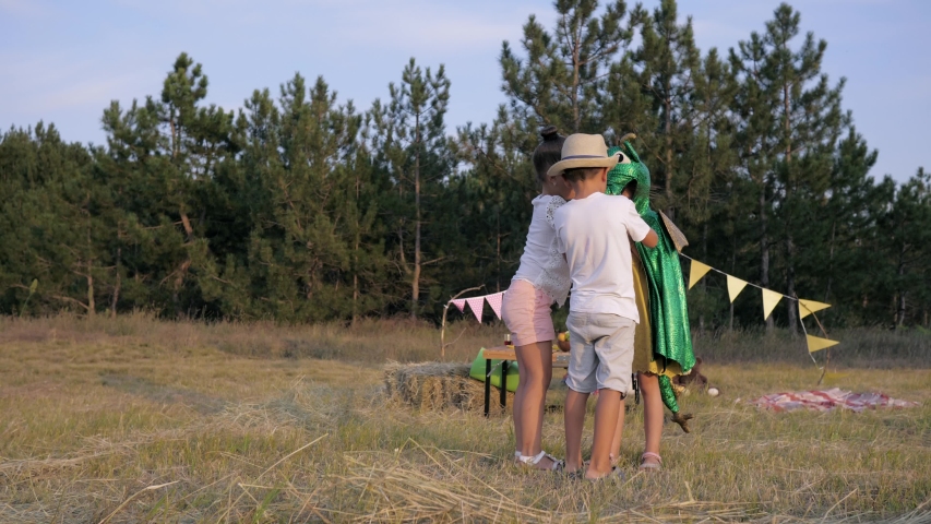 children games, a little female in dragon costume runs with her cheerful friends in the clearing during picnic in forest backdrop of a wigwam